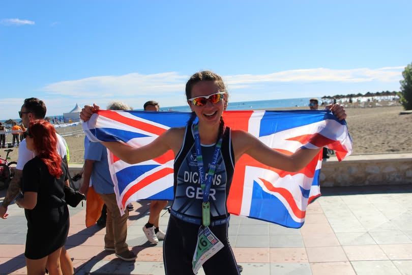 A lady holding the British flag up behind her wearing sunglasses and a tri-suit with the beach and ocean in the background.