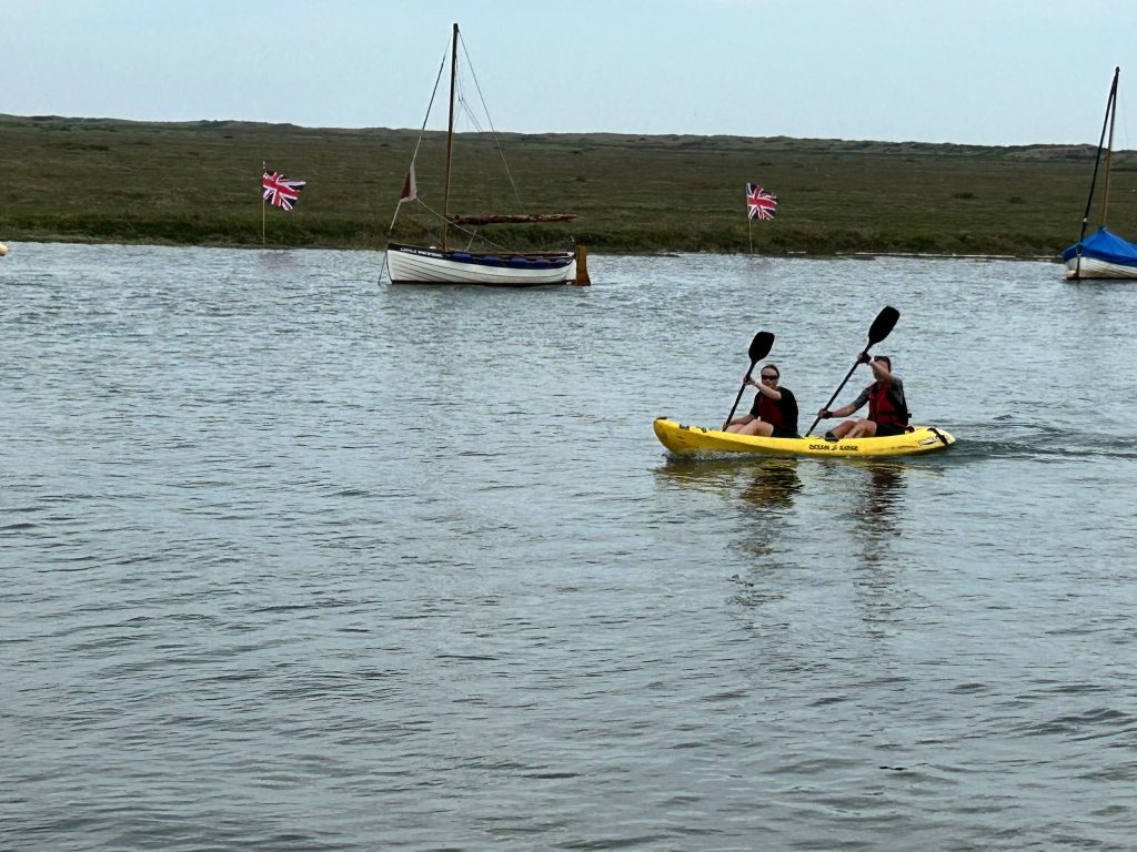 2 ladies in a kayak with 2 boats and 2 British flags in the background placed on a marsh area.