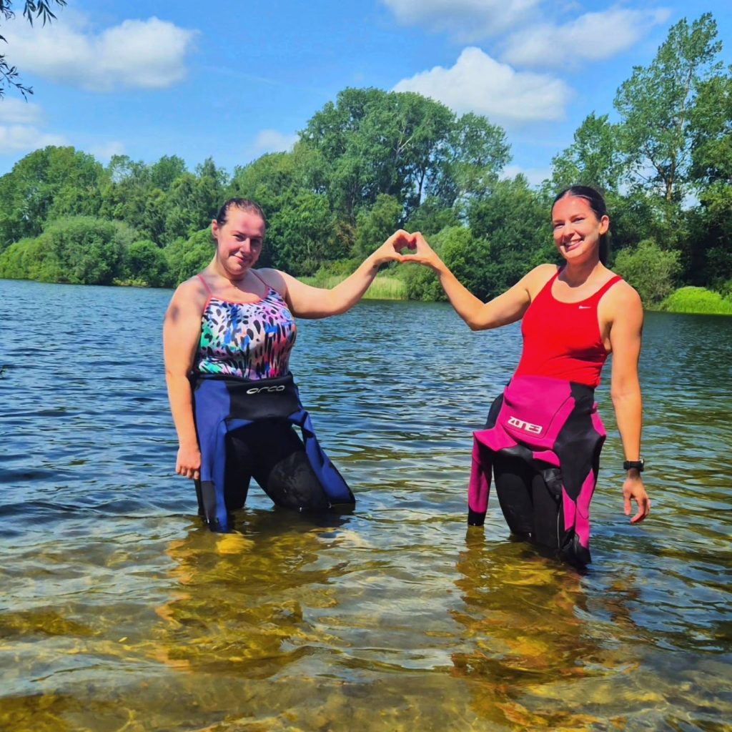2 efficient endurance athletes standing in a lake with wetsuits at their hips crating a love heart with their hands connected.